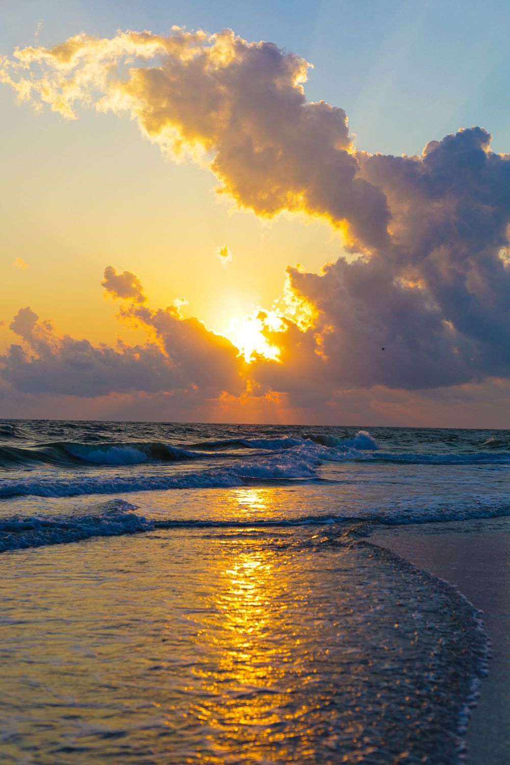 Golden sunrise over ocean waves with glowing clouds reflecting light on a sandy beach.