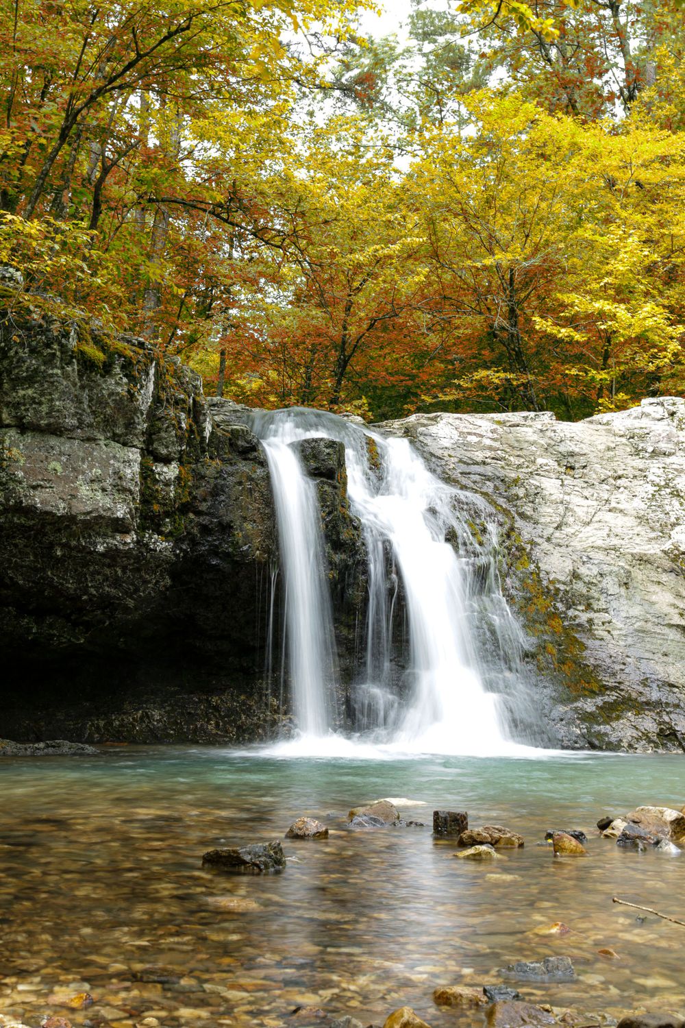 A scenic waterfall cascades over white rocks into a clear pool surrounded by vibrant autumn foliage.