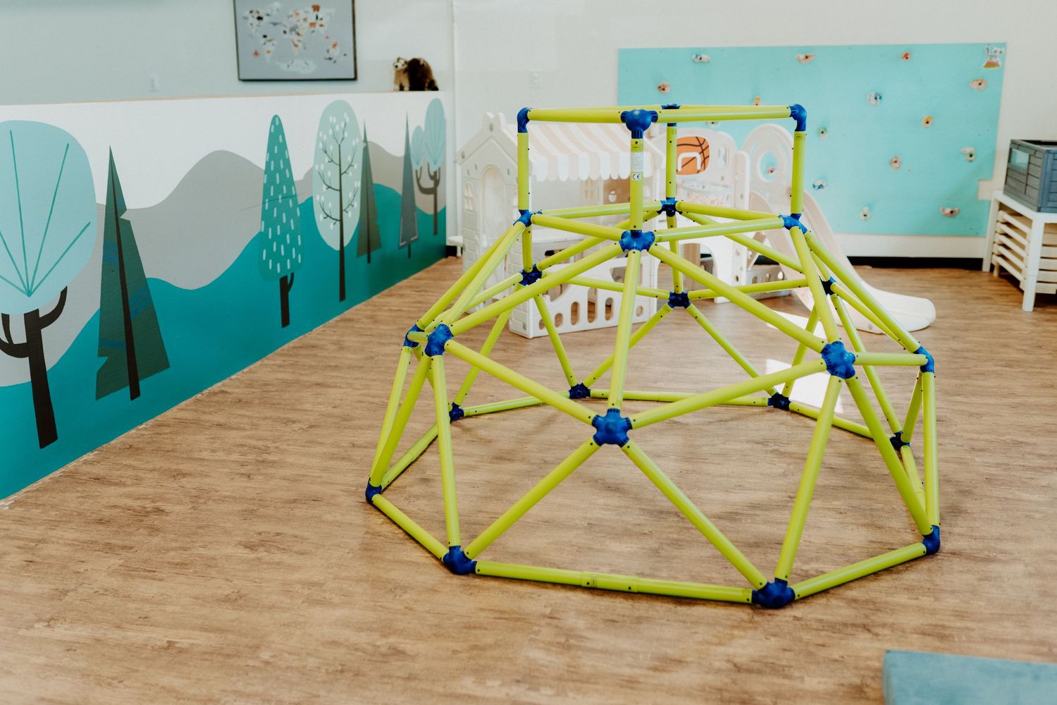 Yellow geometric climbing dome structure in a classroom with blue wall murals and hardwood floors.
