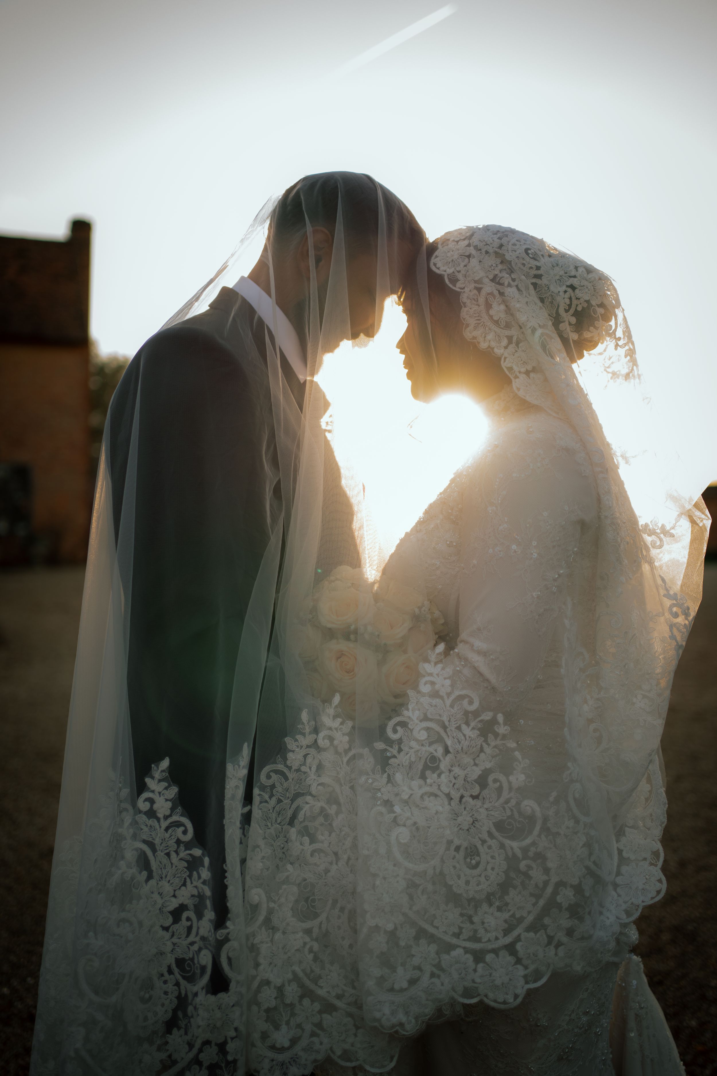 A romantic silhouette of a bride and groom in lace veils touching foreheads at sunset with golden backlight.