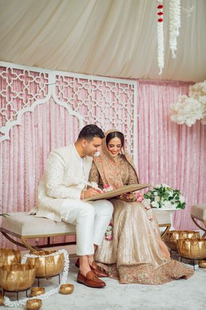 A romantic wedding scene with pink curtains and gold accents features a couple sitting on elegant white and gold furniture.