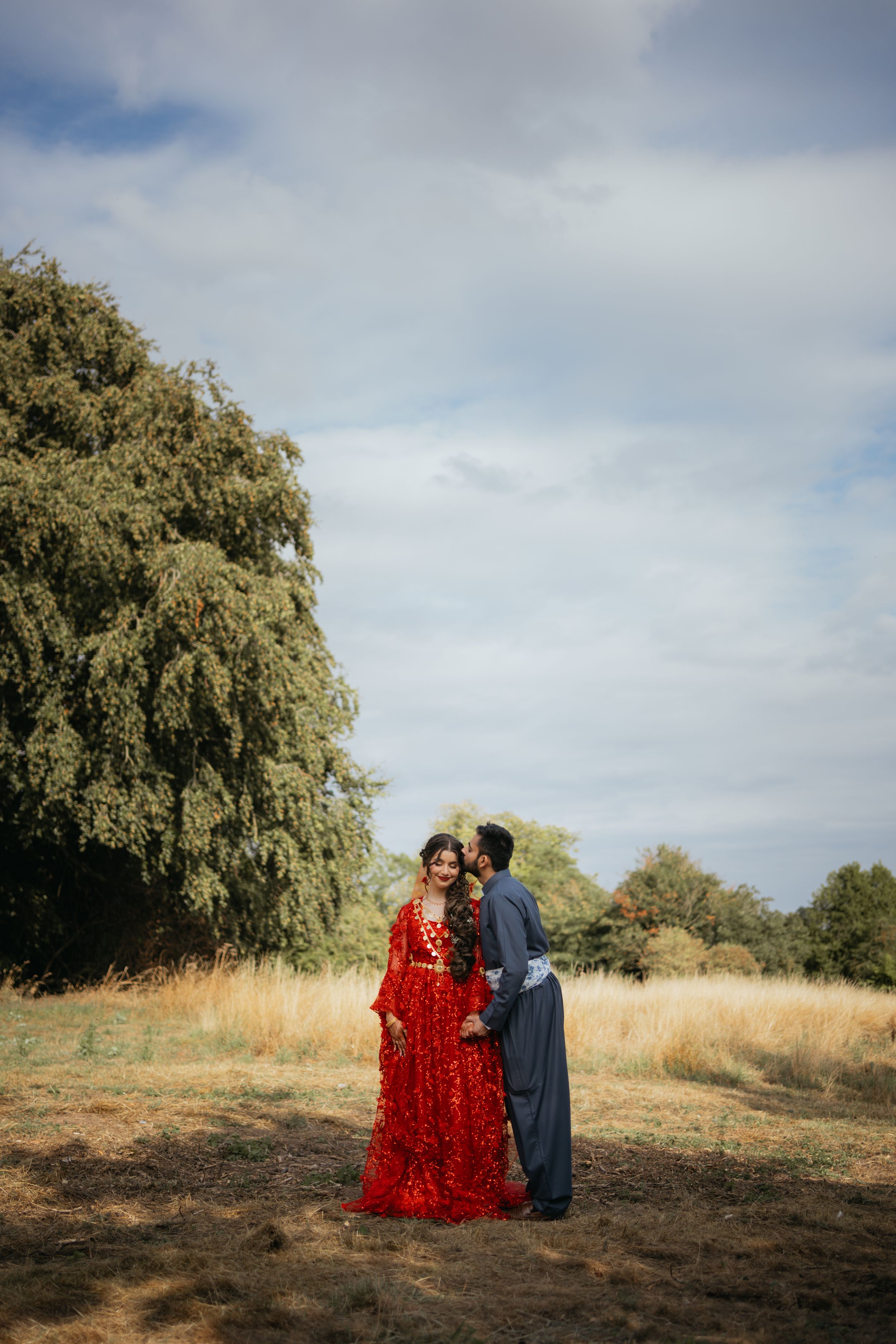 A couple stands together in a scenic field with willow trees, the woman wearing a striking red dress and the man in blue.
