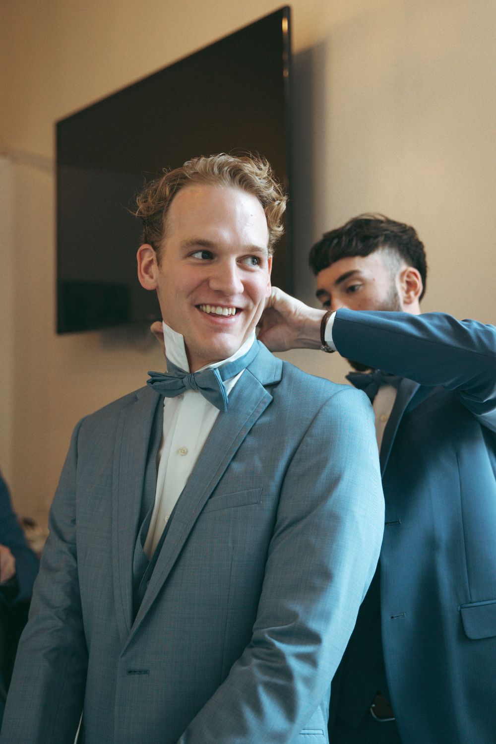 Groomsmen in blue suits help adjust the groom's bowtie before the wedding ceremony.