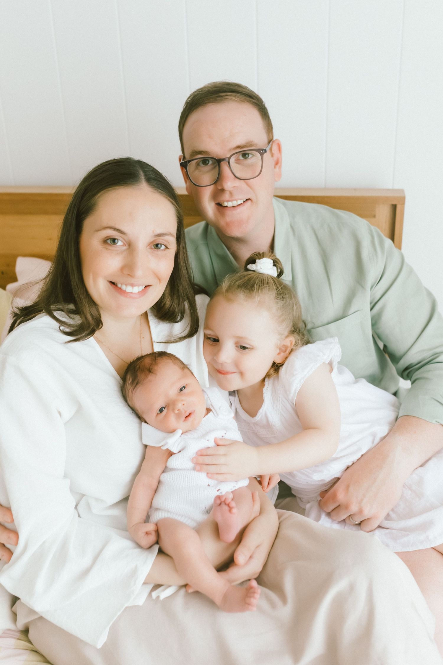 Happy family of four with newborn baby posing together on a bed, smiling for a bright indoor portrait photo.
