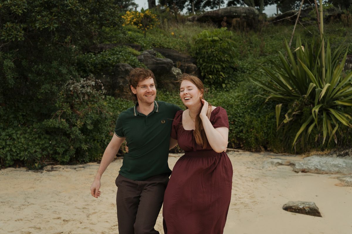 A couple laughing and walking together on a sandy beach surrounded by lush green trees in watsons bay sydney proposal photoshoot