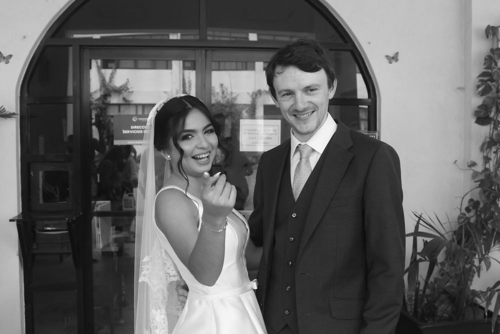 Black and white wedding photo of smiling bride in veil and groom in suit with tie posing together outdoors.