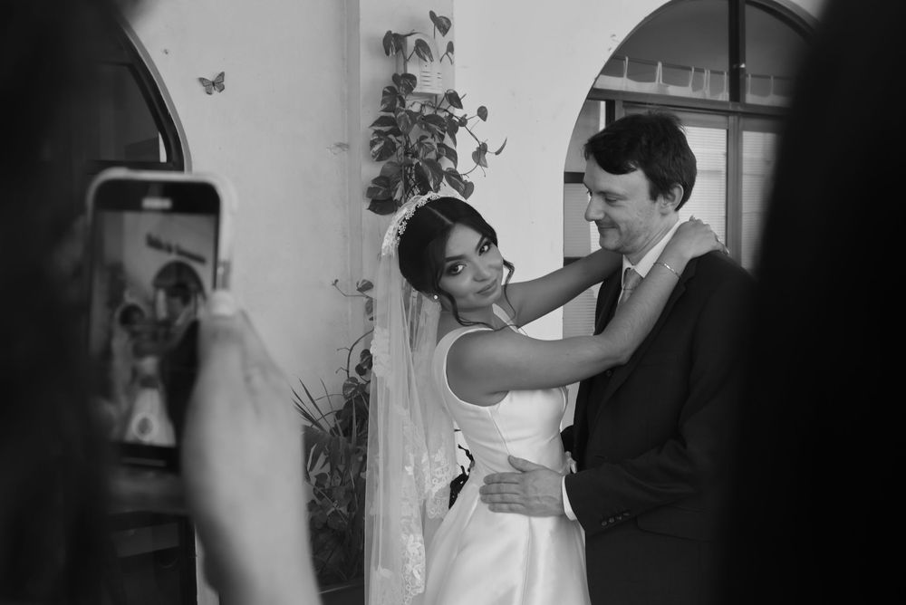 Black and white wedding photo of bride and groom embracing while guest photographs them on smartphone.