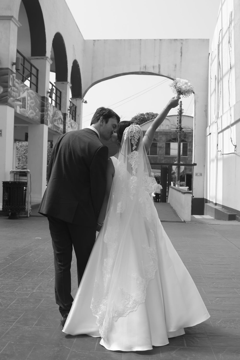 Black and white photo of a bride and groom walking, bride raising bouquet in celebration under an archway.