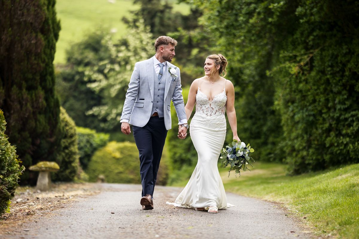 Bride and groom walking together on a tree-lined path in formal wedding attire at an outdoor garden venue.