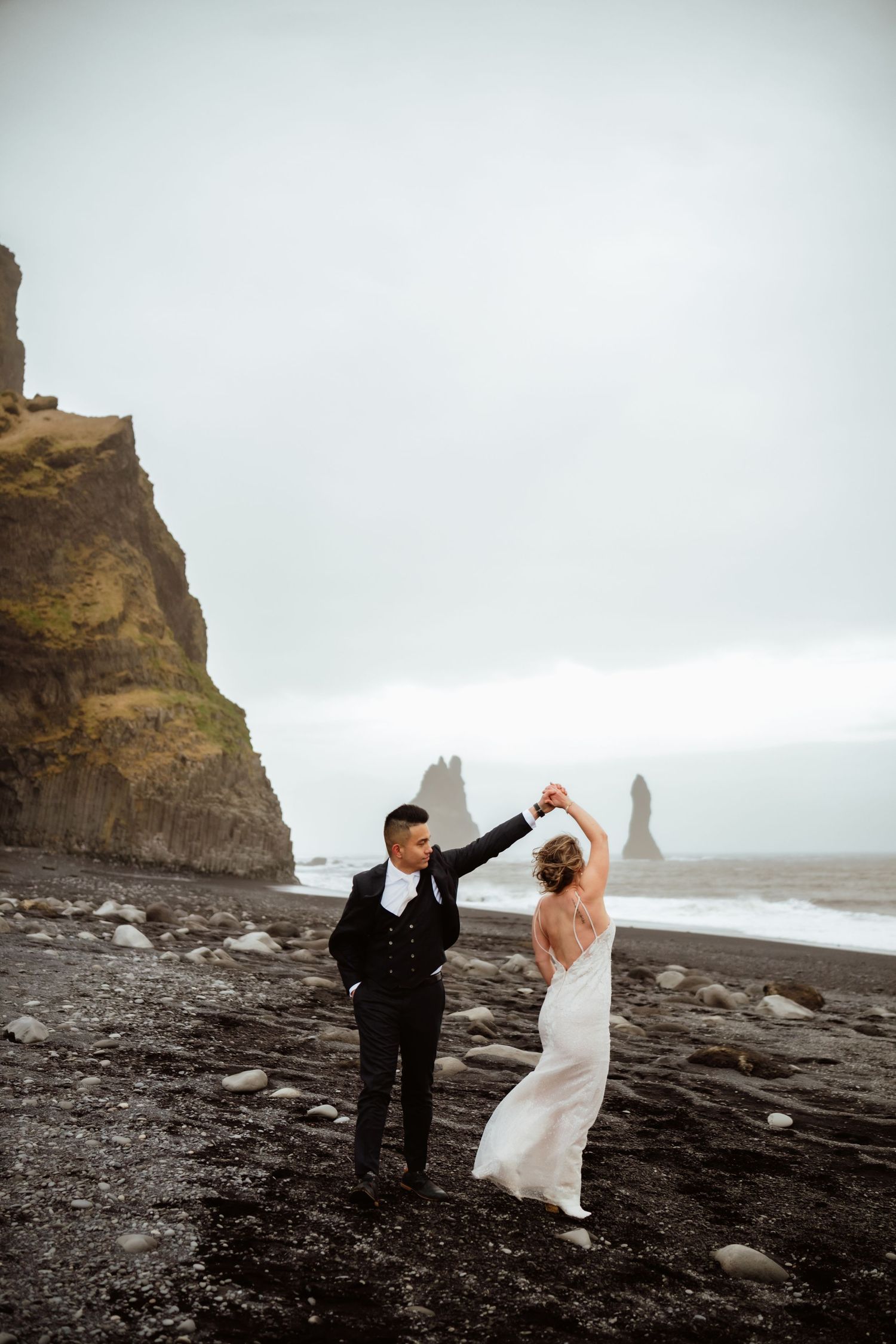 Bride and groom dancing on black sand beach in Iceland with dramatic sea stacks and cliffs in background.