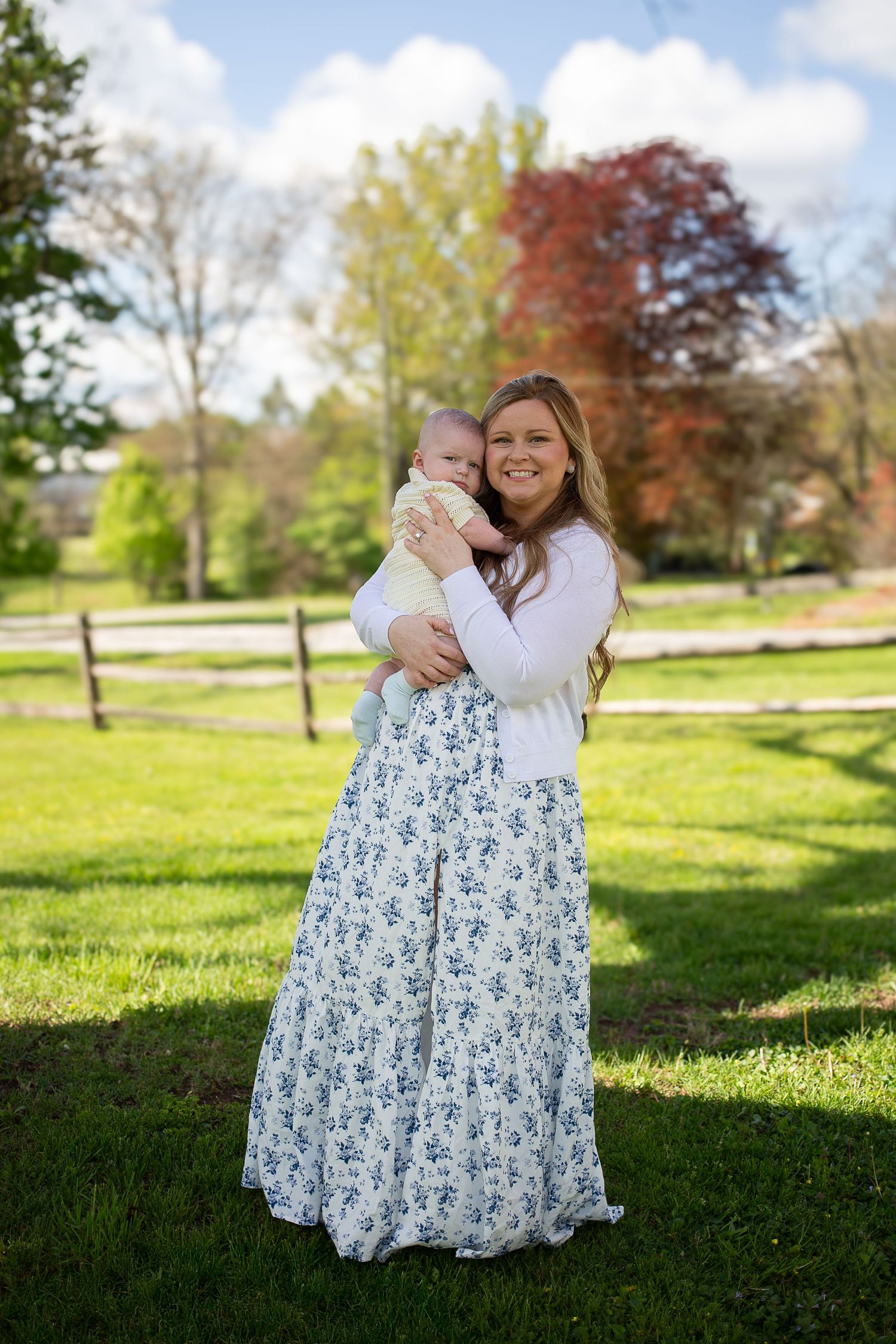 Smiling woman in floral maxi skirt holding a baby outdoors on green grass with trees and fence in background.