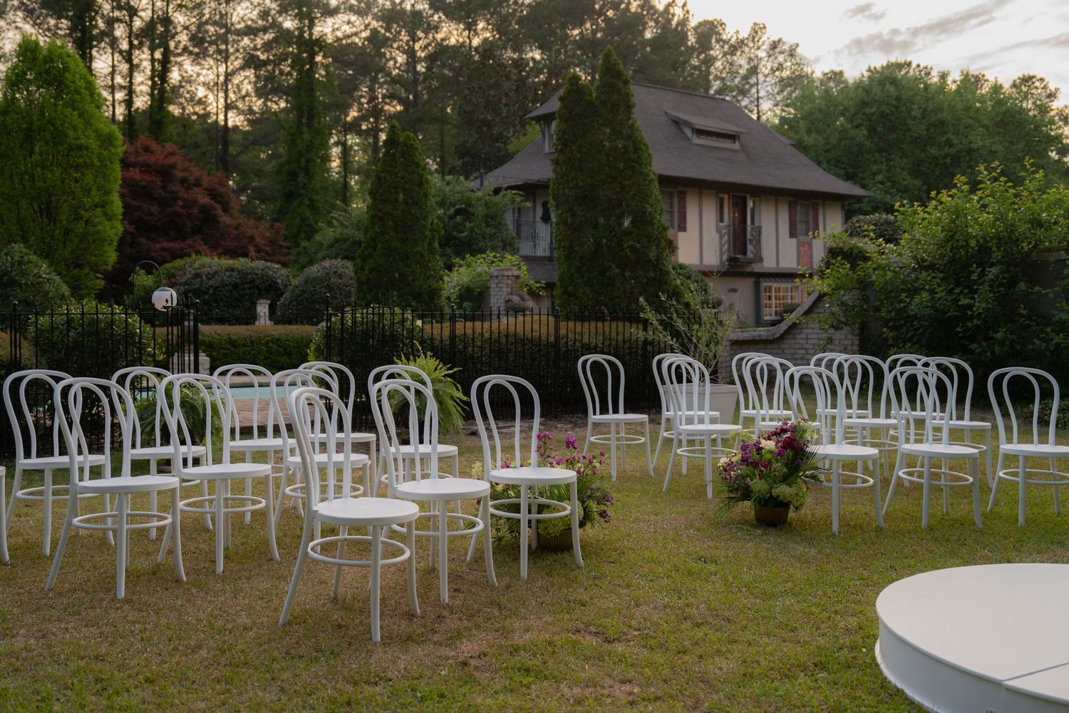 White chairs arranged in rows on a garden lawn for an outdoor wedding ceremony with a house and trees in the background.