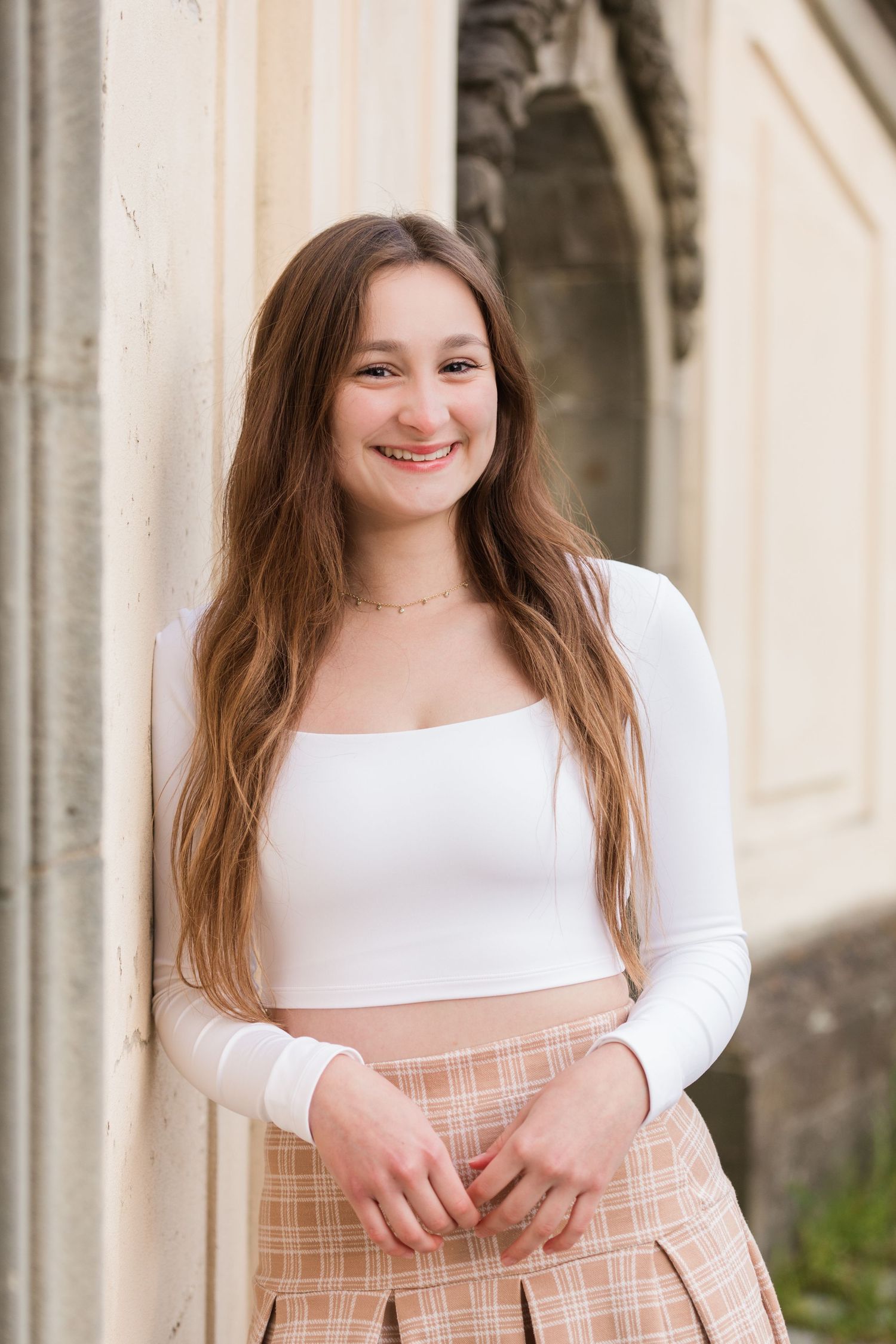 Young woman with long brown hair in white top smiling, resting chin on hand against a stone column.