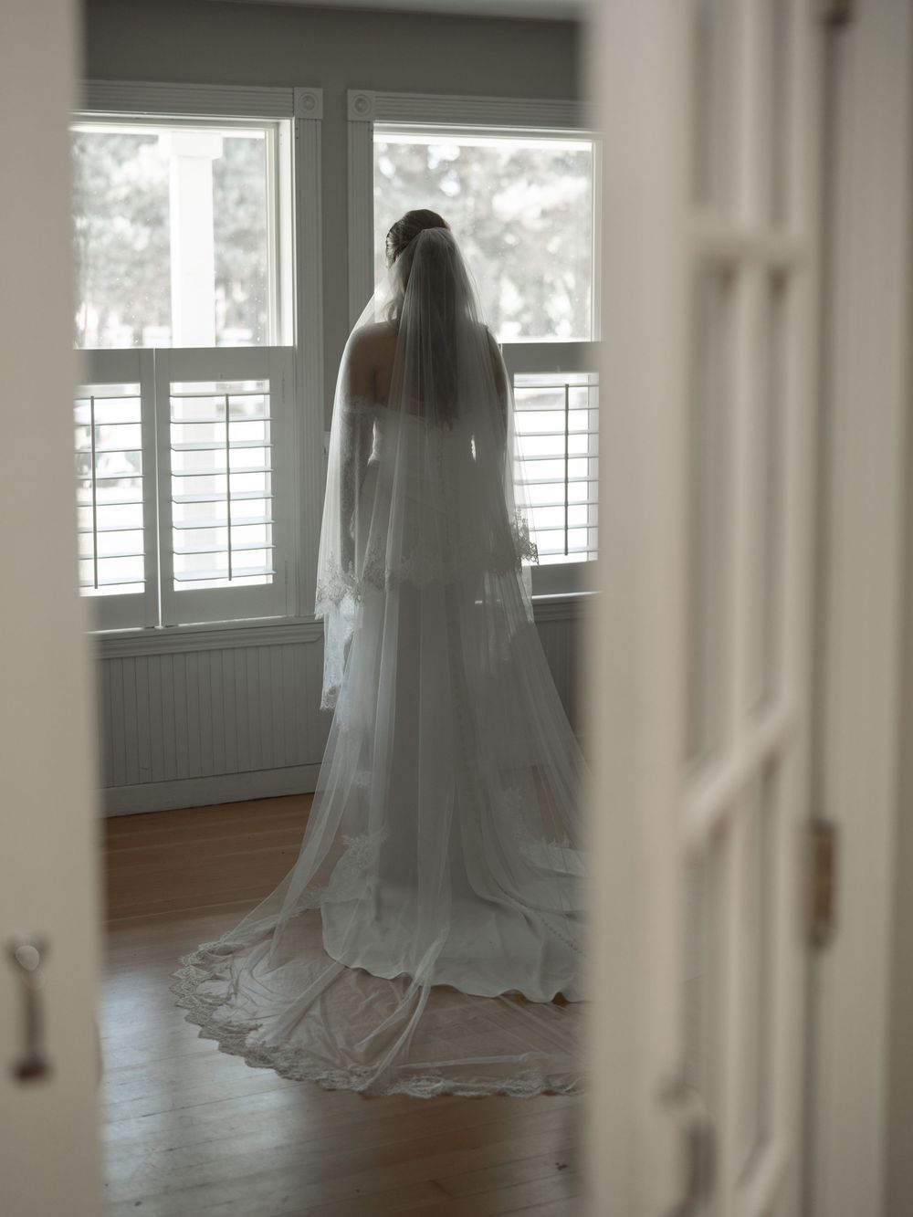 Bride in a flowing white wedding dress and long veil standing by a bright window in a softly lit room.