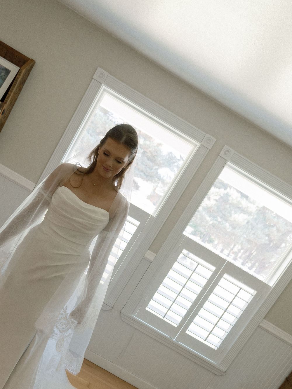 Bride in a strapless white wedding gown with sheer sleeves stands near bright windows in an elegant room.