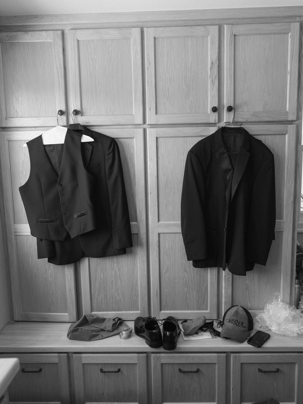 Black and white photo of two dark suits hanging on wooden cabinet doors with shoes and accessories below.