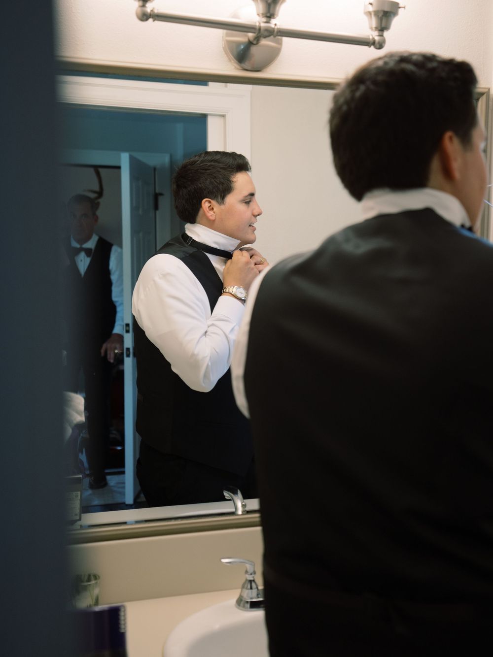 Groom adjusting his bow tie in bathroom mirror while getting ready for wedding day.