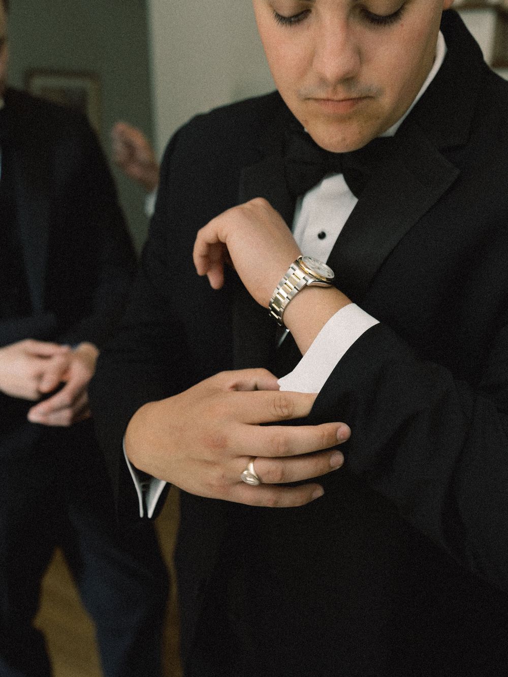 Groom in black tuxedo adjusting cufflinks, wearing a silver watch and ring, preparing for wedding ceremony.