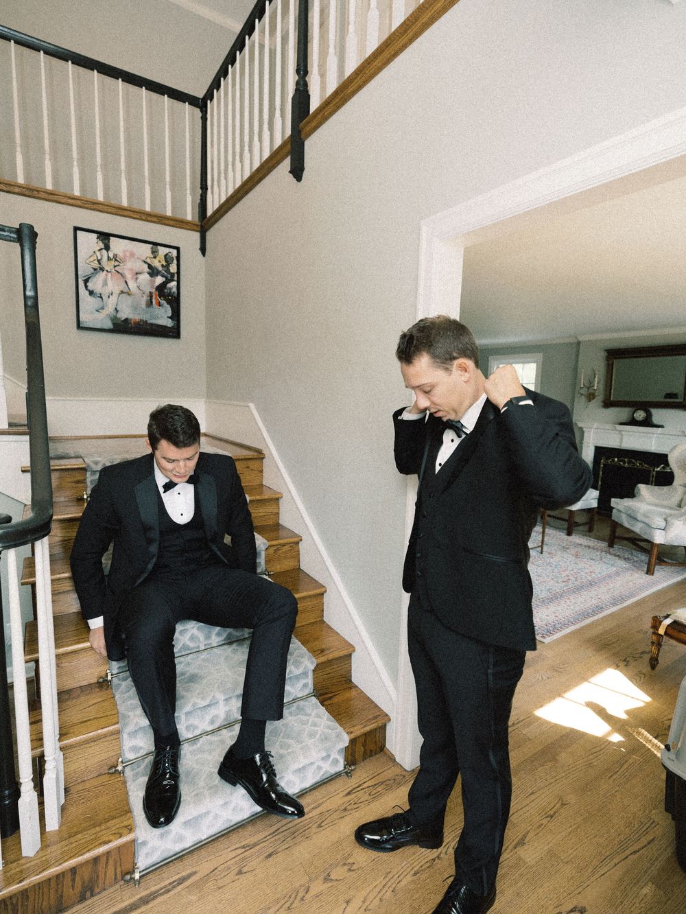Two grooms in black tuxedos prepare for their wedding ceremony on a staircase in an elegant home interior.