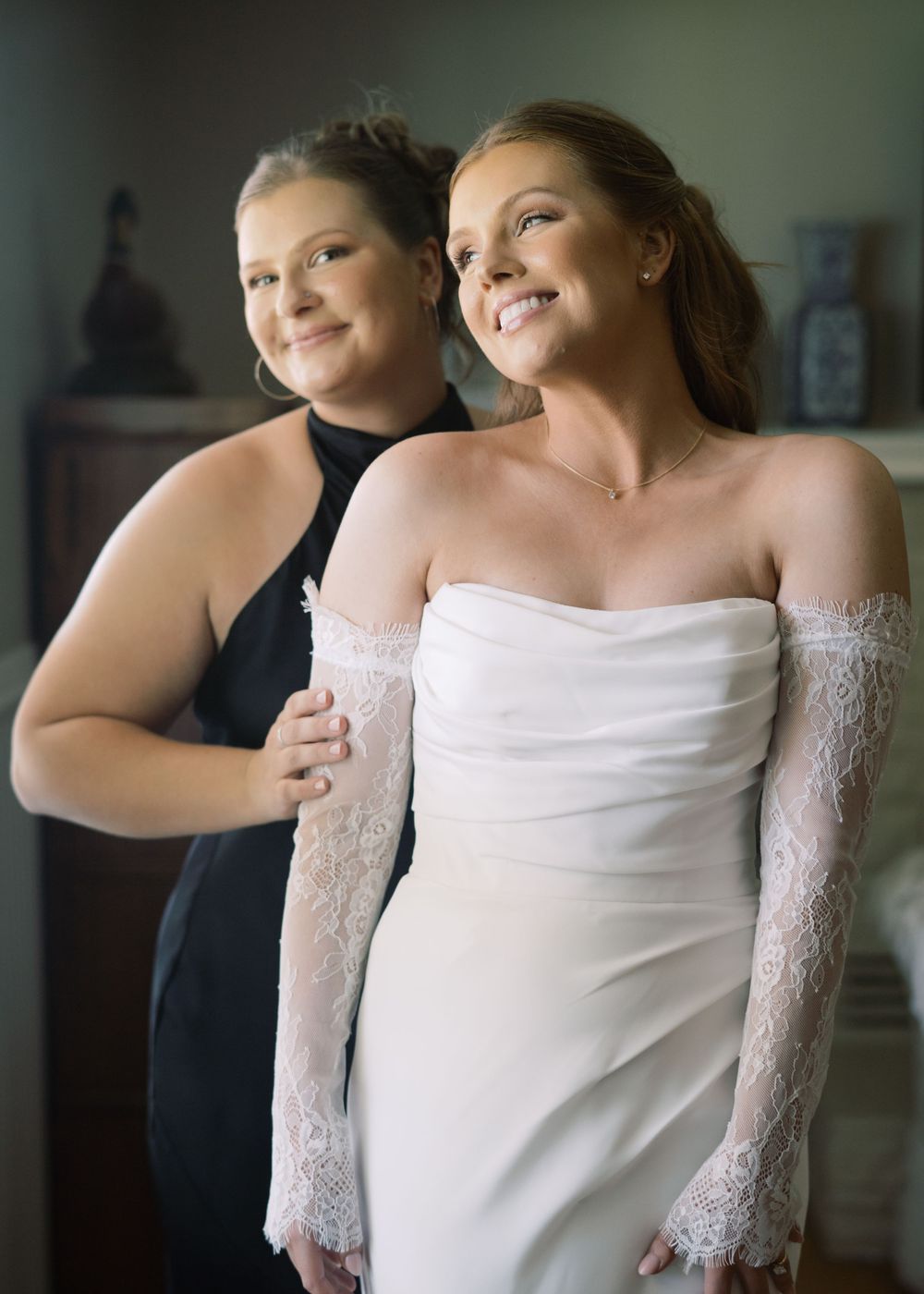 Bride in white strapless gown with lace gloves stands with bridesmaid in black dress during wedding preparations.