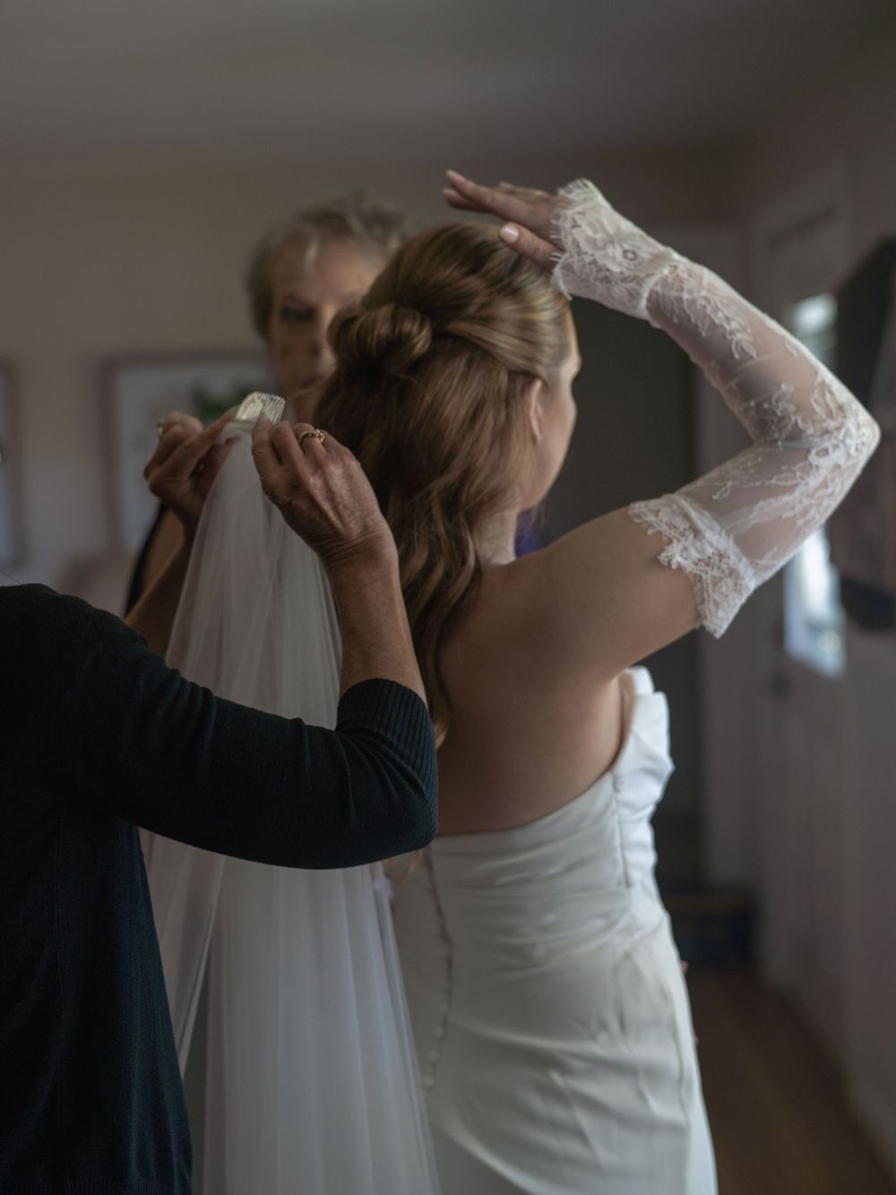 A bride in a white gown and lace gloves has her veil pinned by a helper while viewing herself in a mirror.