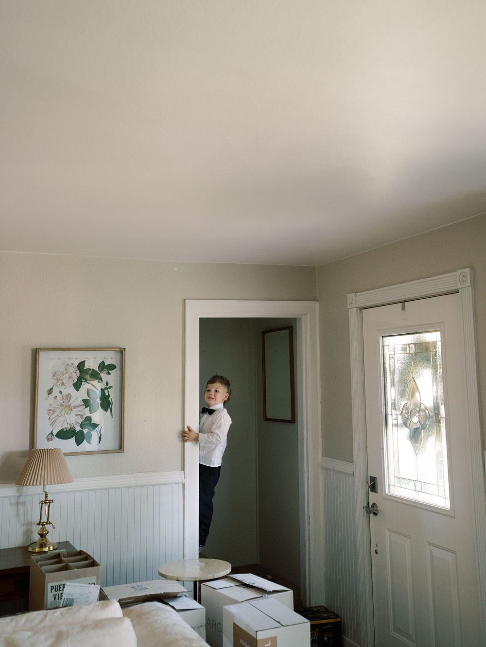 A young boy climbs on a doorframe inside a home with moving boxes, a lamp, and botanical wall art nearby.
