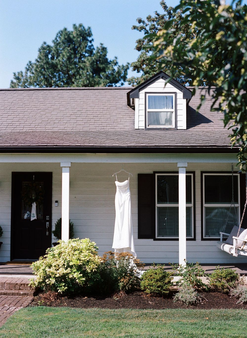 White farmhouse with black shutters displays a hanging wedding dress on the front porch, surrounded by lush greenery.