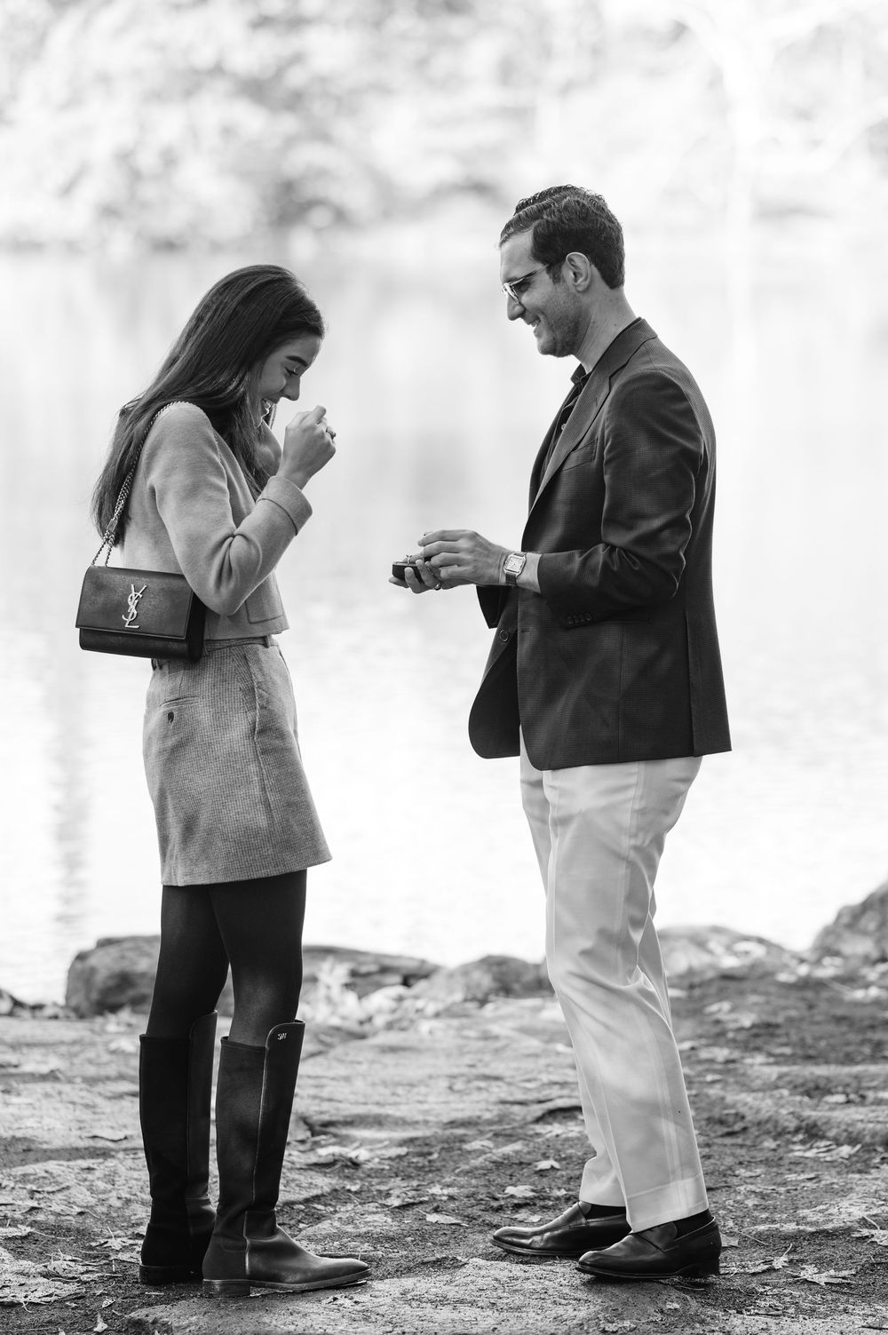 Black and white photo of a man presenting an engagement ring to an emotional woman near a lake.