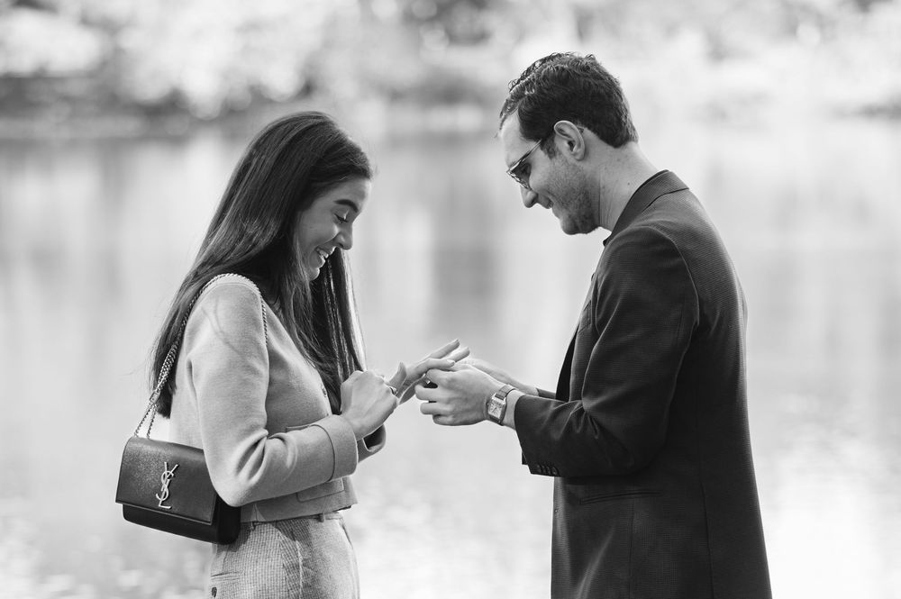 Black and white photo of a man placing an engagement ring on a woman's finger by a lake.