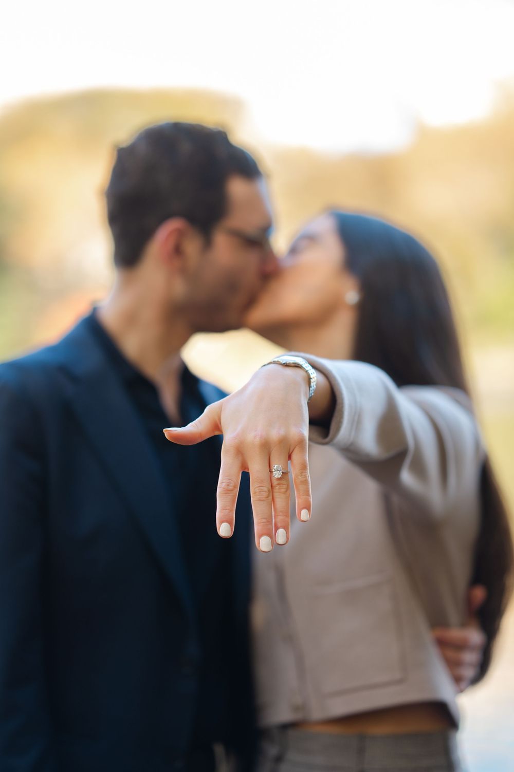 Couple kissing while woman extends hand showing engagement ring outdoors in warm golden light.