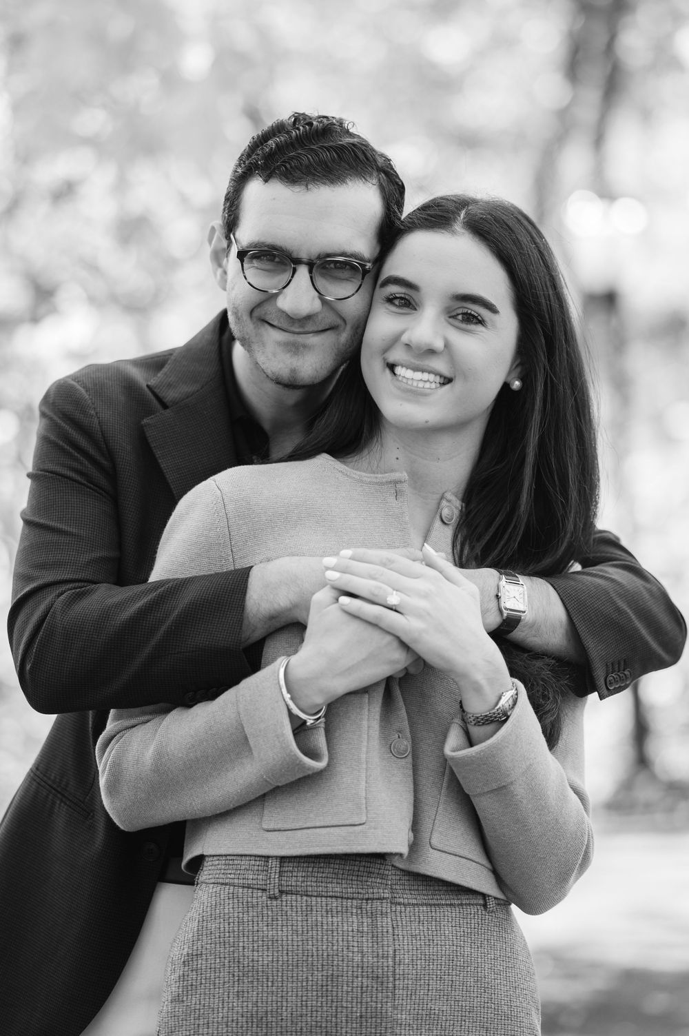 Black and white photo of smiling couple embracing outdoors, woman in beige suit, man in glasses hugging her from behind.