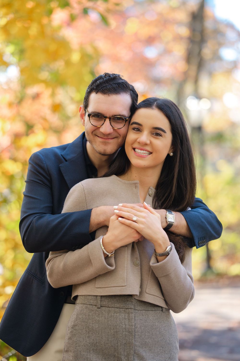 Couple embracing in autumn park, woman in beige suit smiling, man in navy blazer and glasses hugging her from behind.
