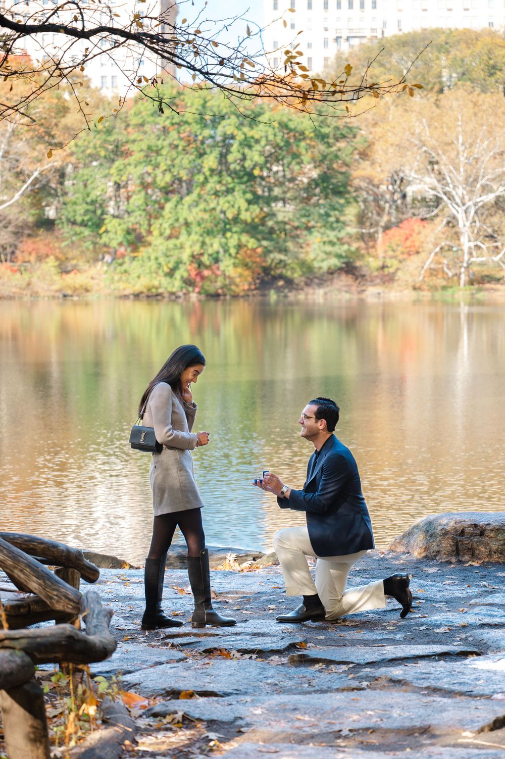 Man proposes to woman on one knee by a lake in Central Park during autumn.