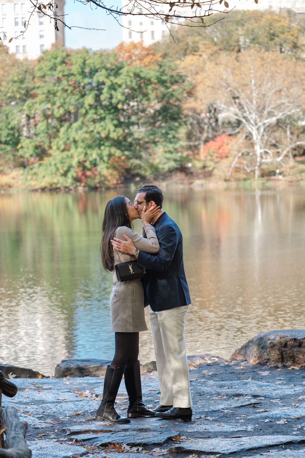 Couple sharing a romantic kiss by a serene lake in Central Park during autumn, surrounded by colorful foliage.