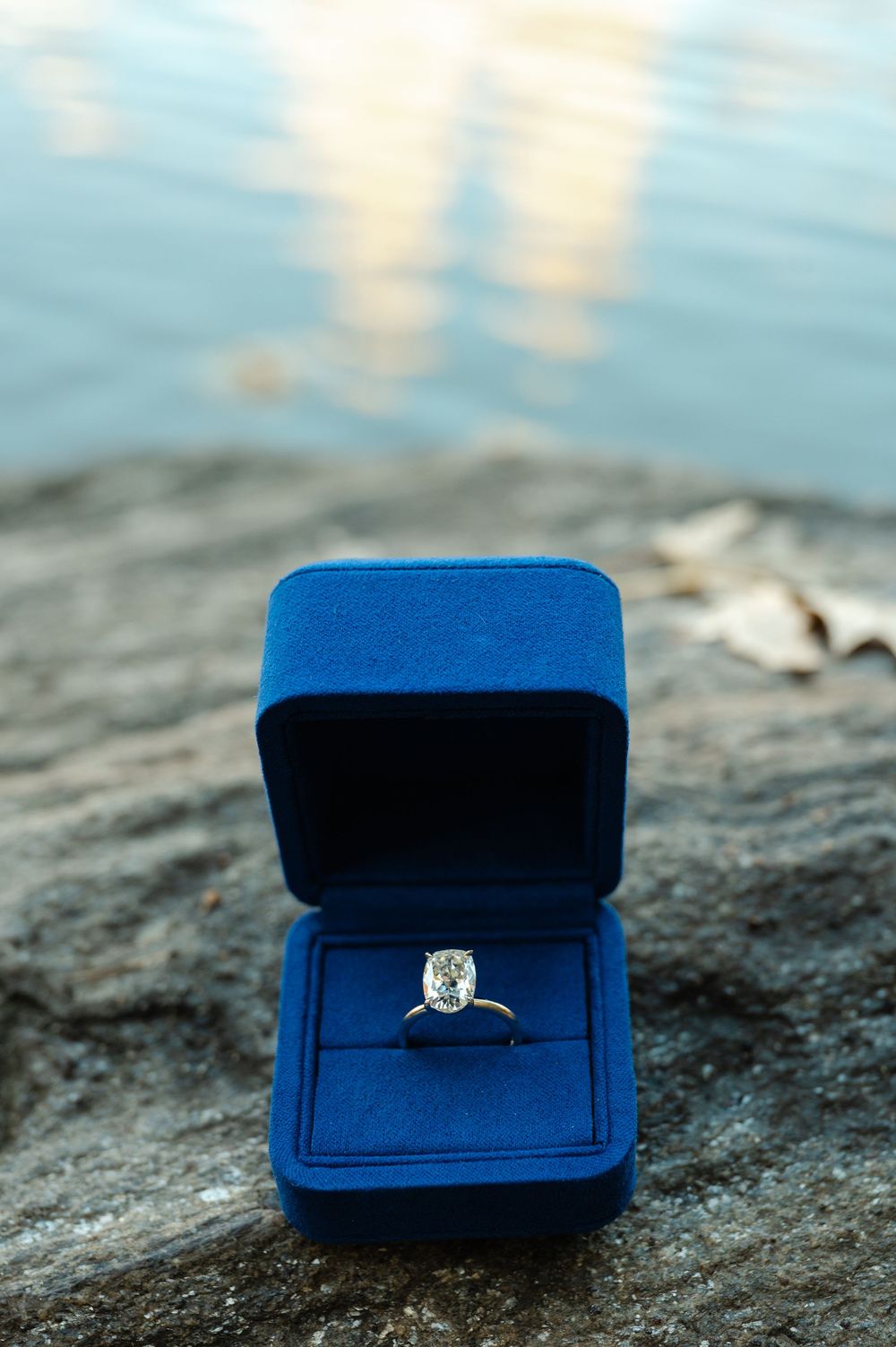 Diamond solitaire engagement ring in a blue velvet box on a rock by water.