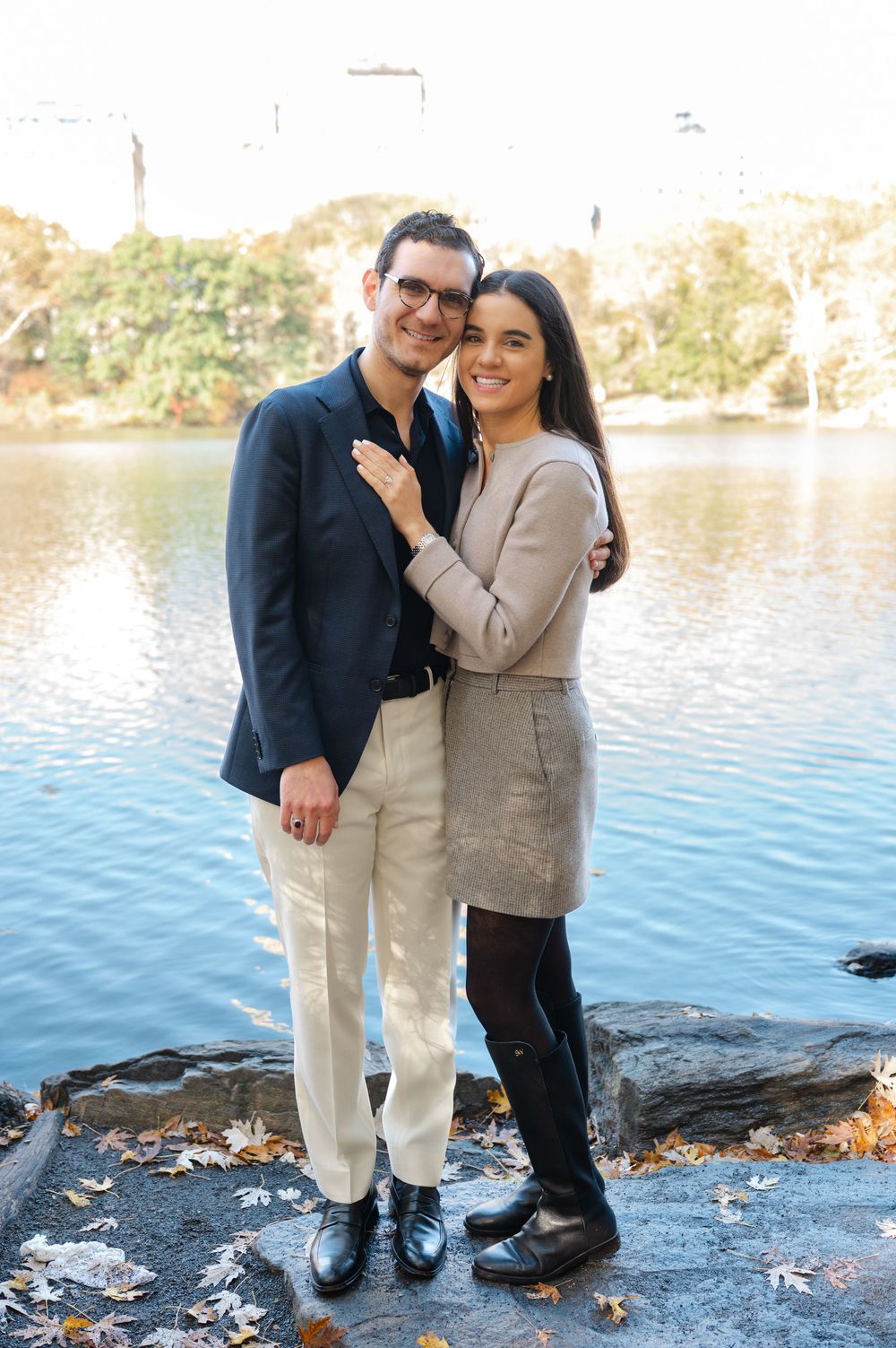 A couple poses romantically by a lakeside in Central Park during autumn, the man in a navy blazer and the woman in a beige dress.