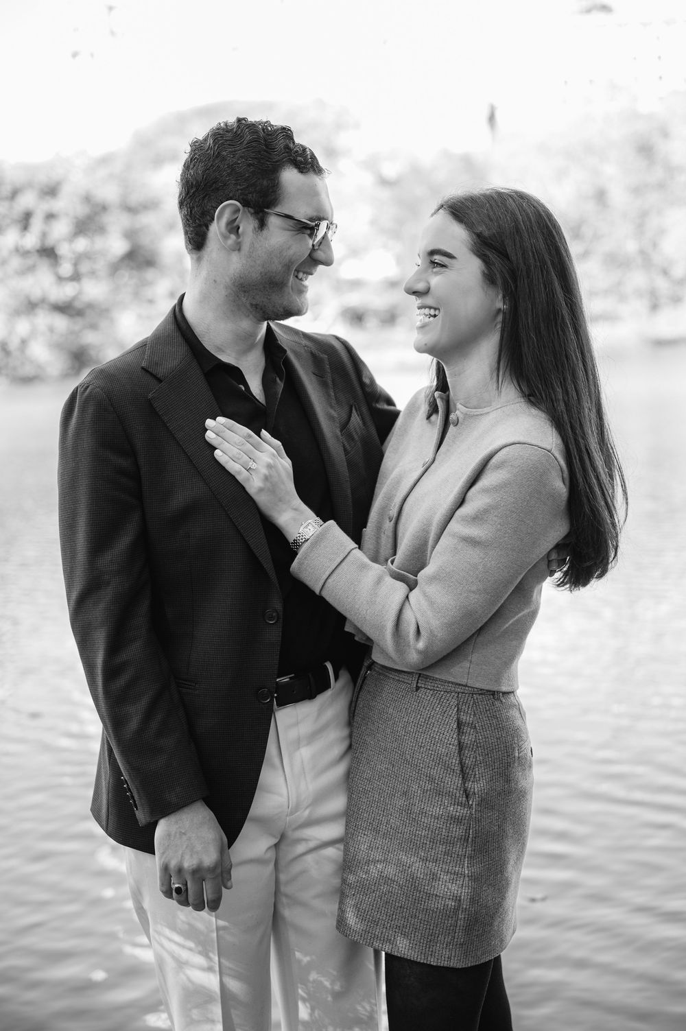 Black and white photo of a smiling couple gazing at each other near a waterfront, woman placing hand on mans chest.