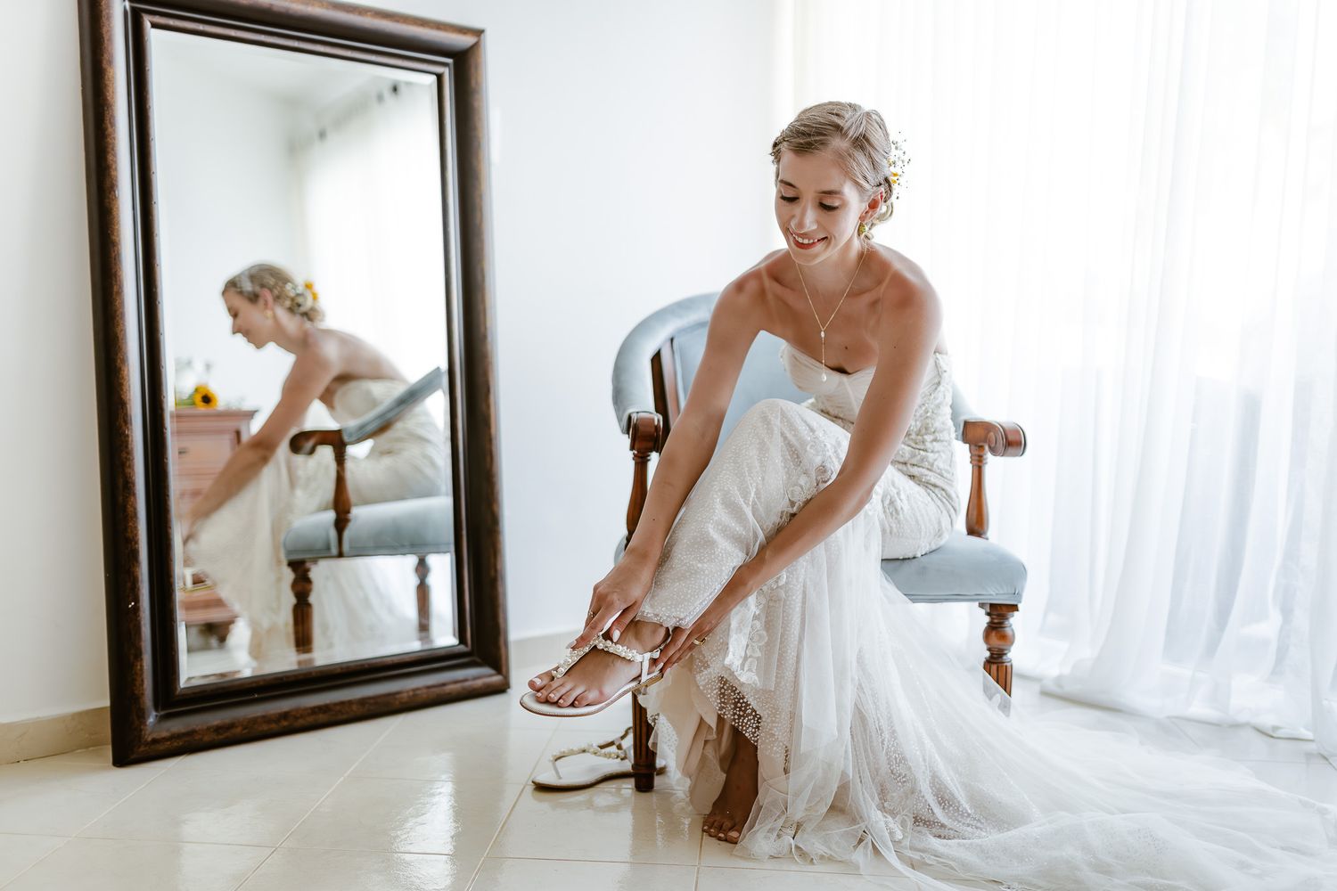 A bride in a white wedding dress adjusts her shoe while sitting in front of a mirror in a sunlit room.