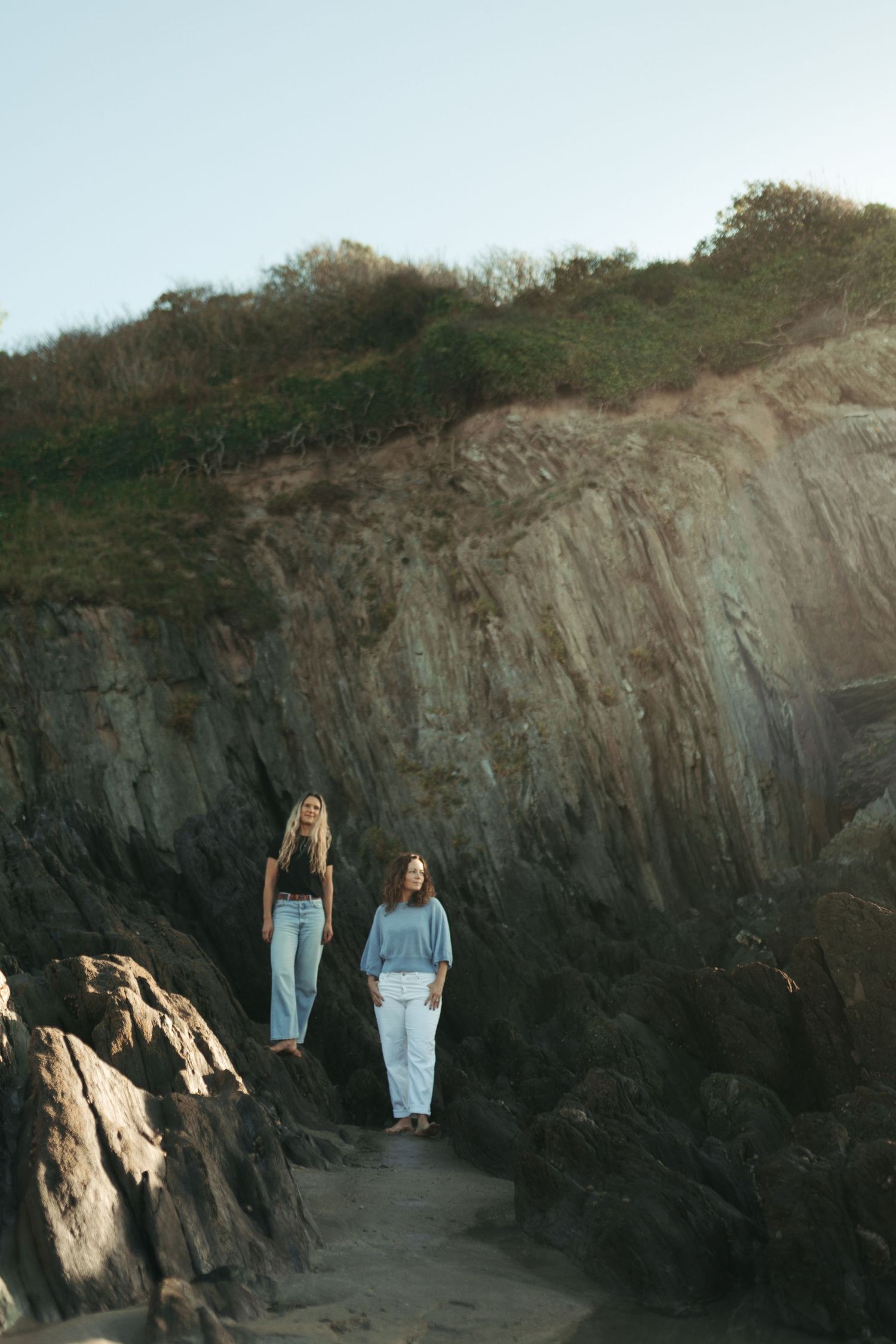 Two women in casual outfits stand on rocky coastal cliffs with green vegetation above them.