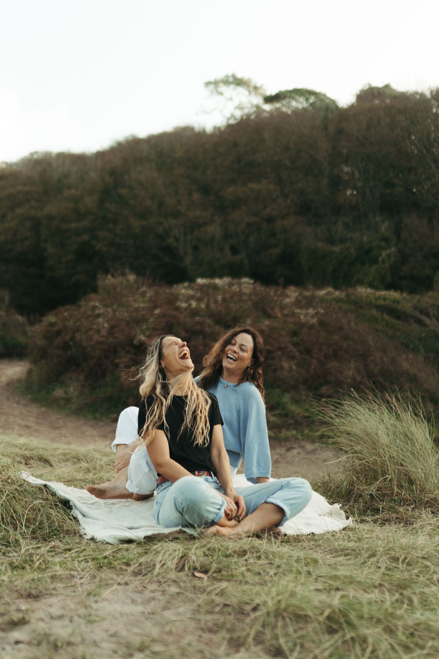 Two women sitting on a blanket in a grassy field, laughing joyfully with autumn shrubs in the background.