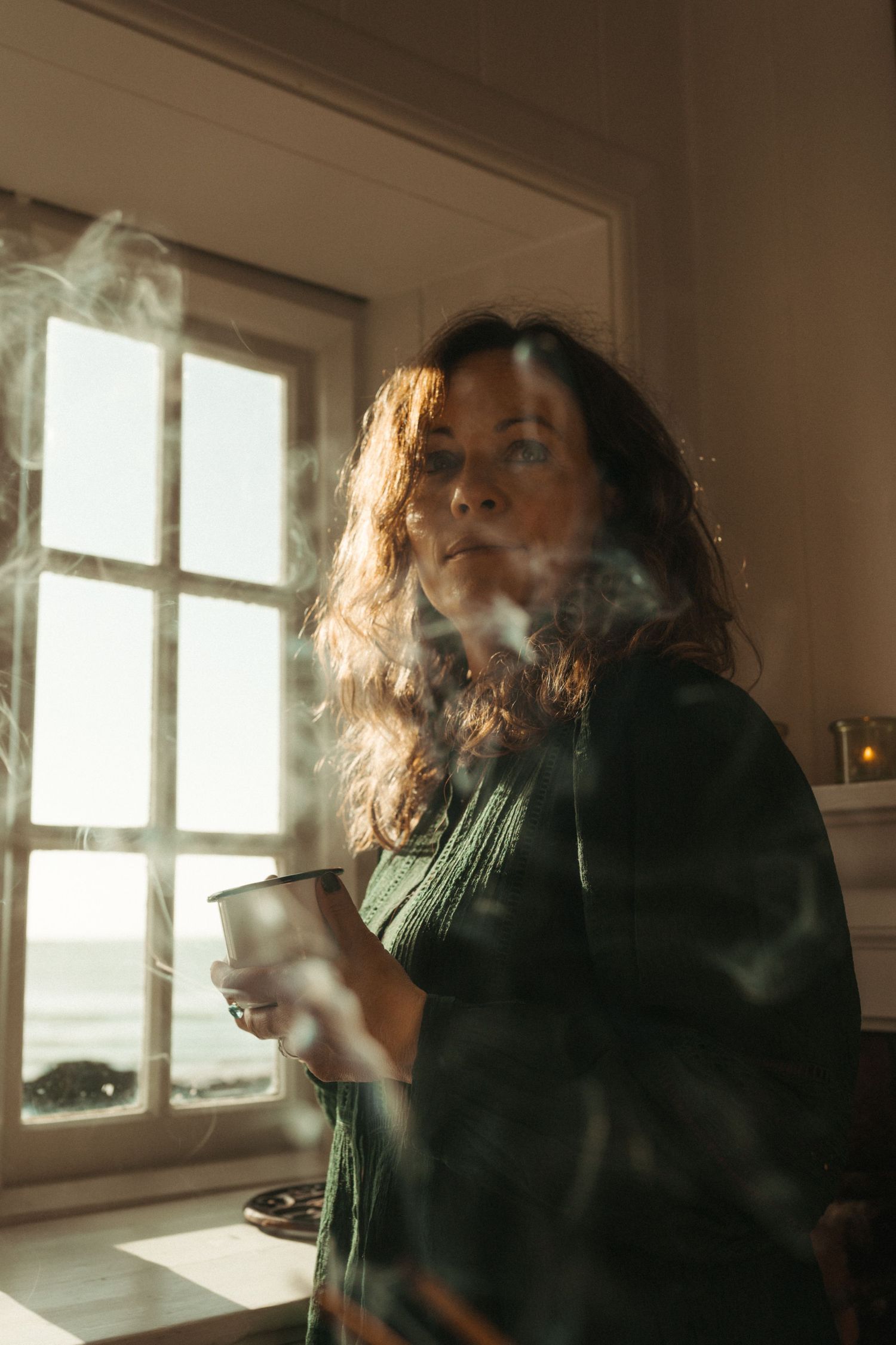 Woman holding a steaming mug by a sunlit window with ocean view, wrapped in a green sweater.