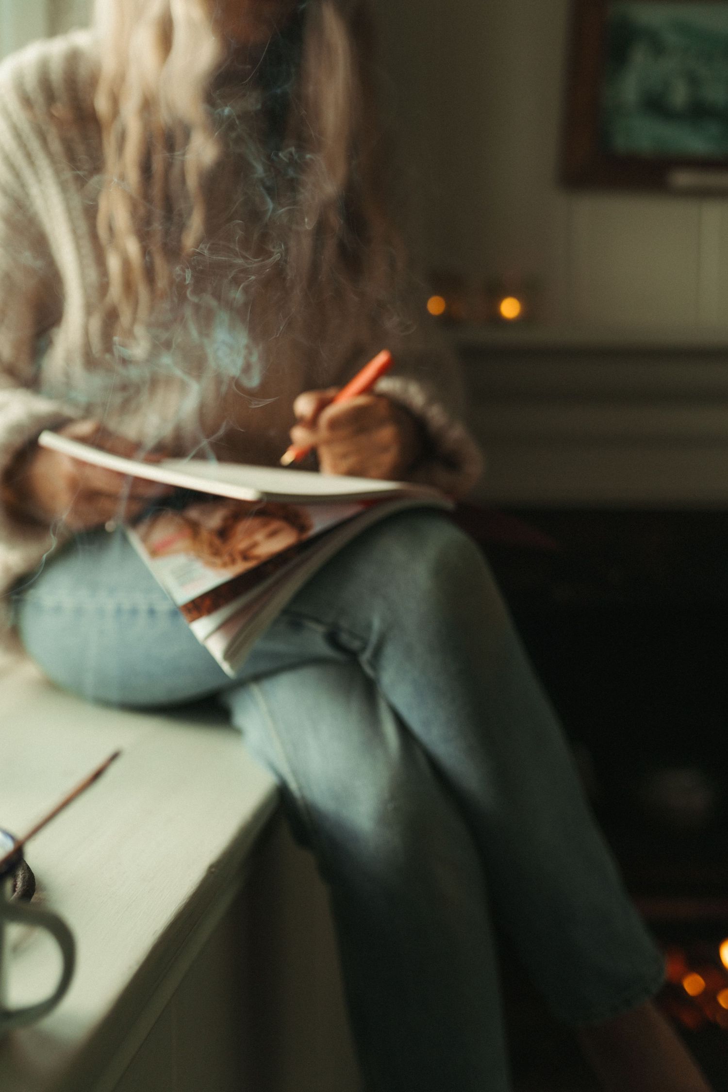 Woman in cozy sweater and jeans sitting on windowsill writing in notebook with orange pencil in warm light.