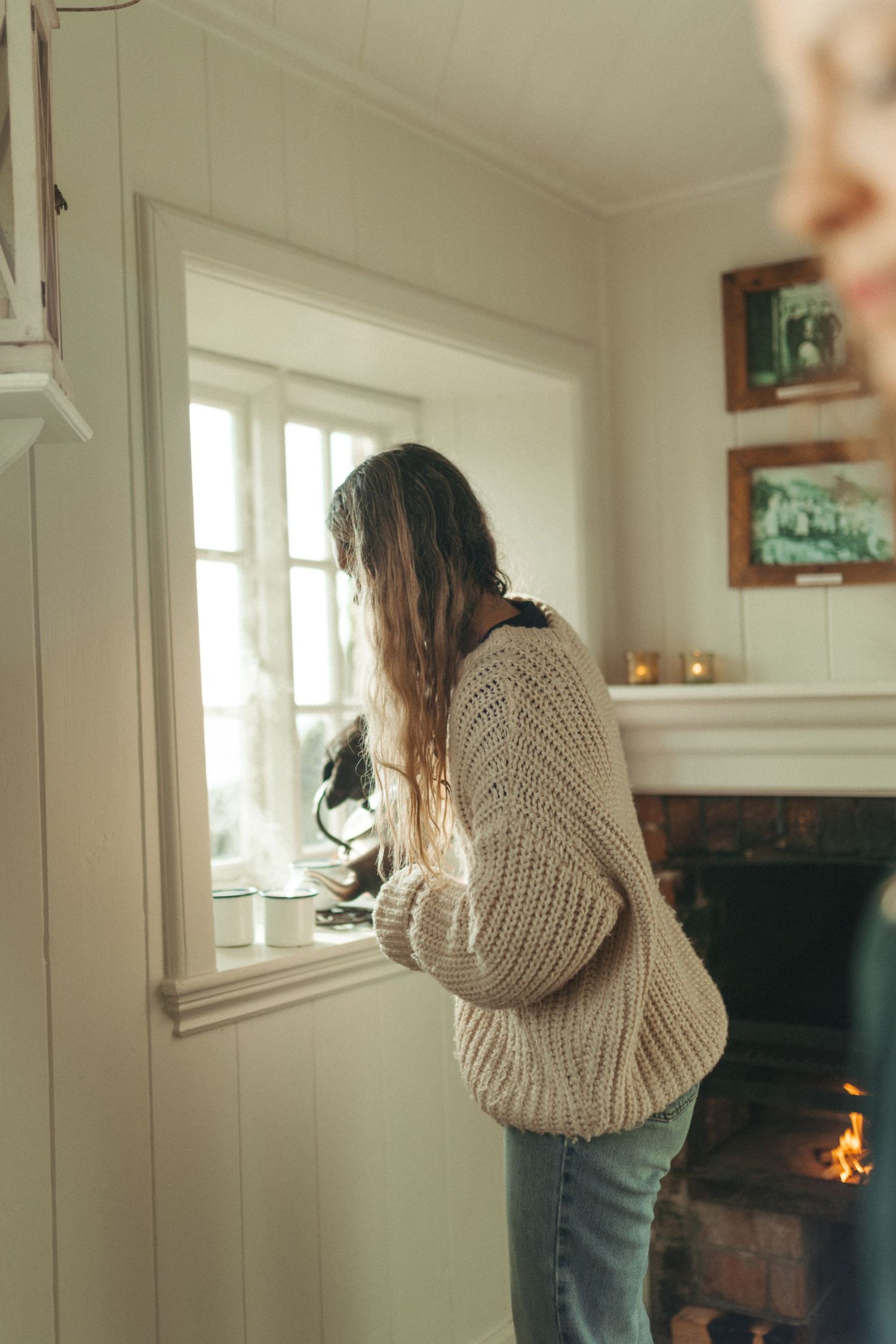 Woman in chunky knit sweater and jeans stands by window in cozy cottage with fireplace.