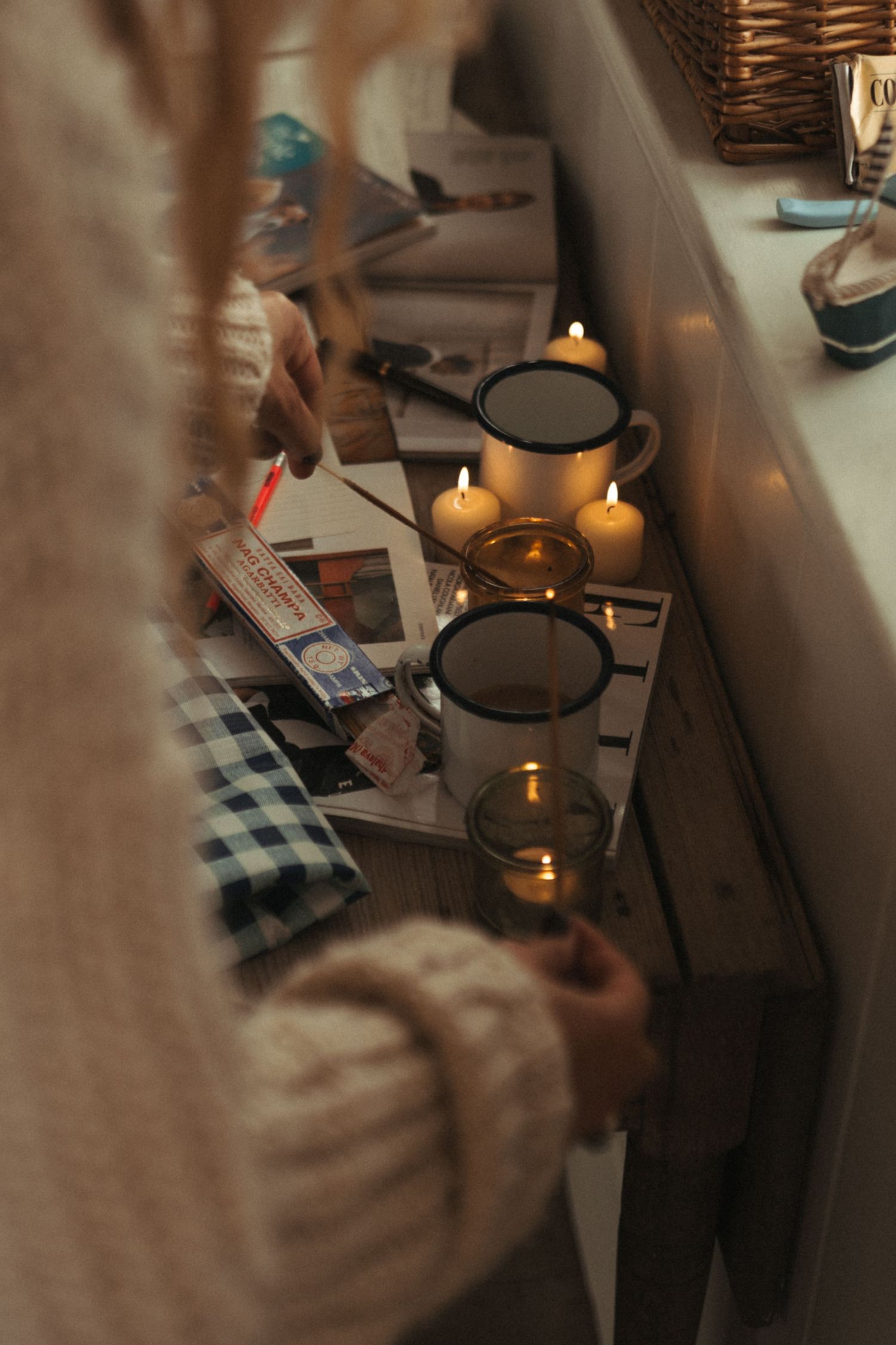 Person in cozy knit sweater lighting candles in enamel mugs on a wooden shelf with scattered magazines.