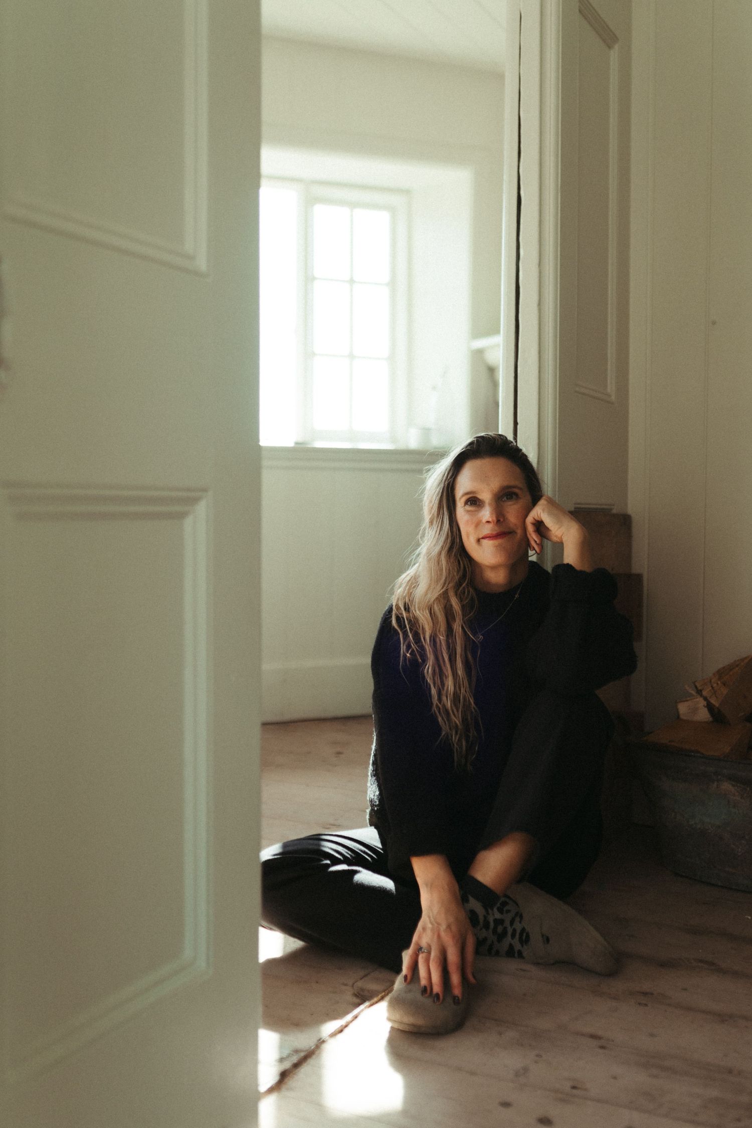 Woman with long hair sitting on wooden floor leaning against wall in bright sunlit hallway.