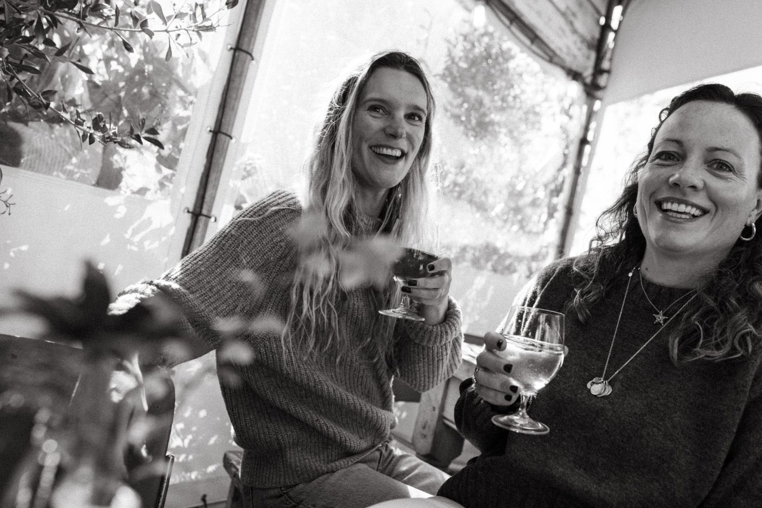 Two women laughing and holding wine glasses in a cozy greenhouse setting, black and white photo.