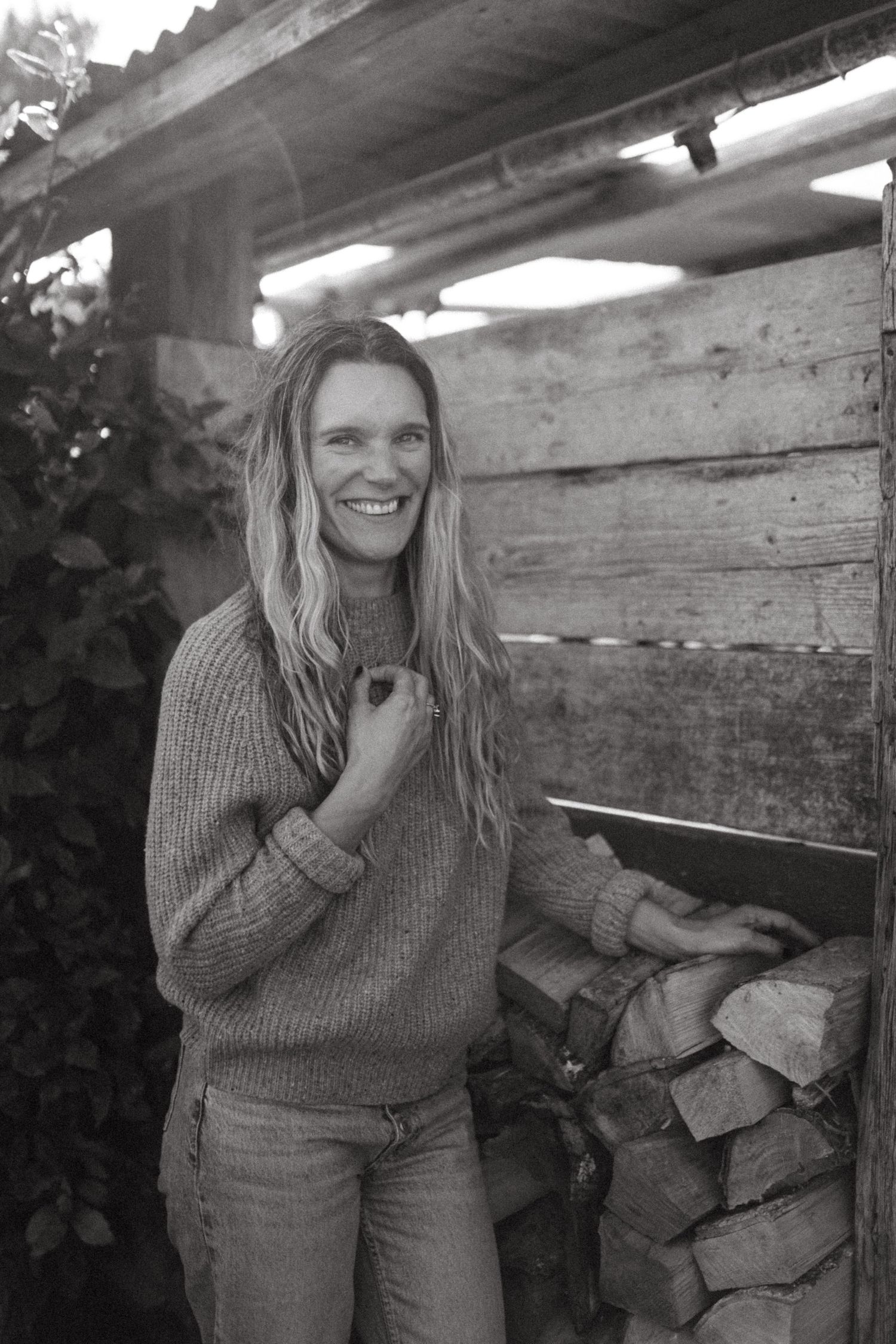 Smiling woman in knit sweater and jeans standing beside stacked firewood in a rustic wooden shed.