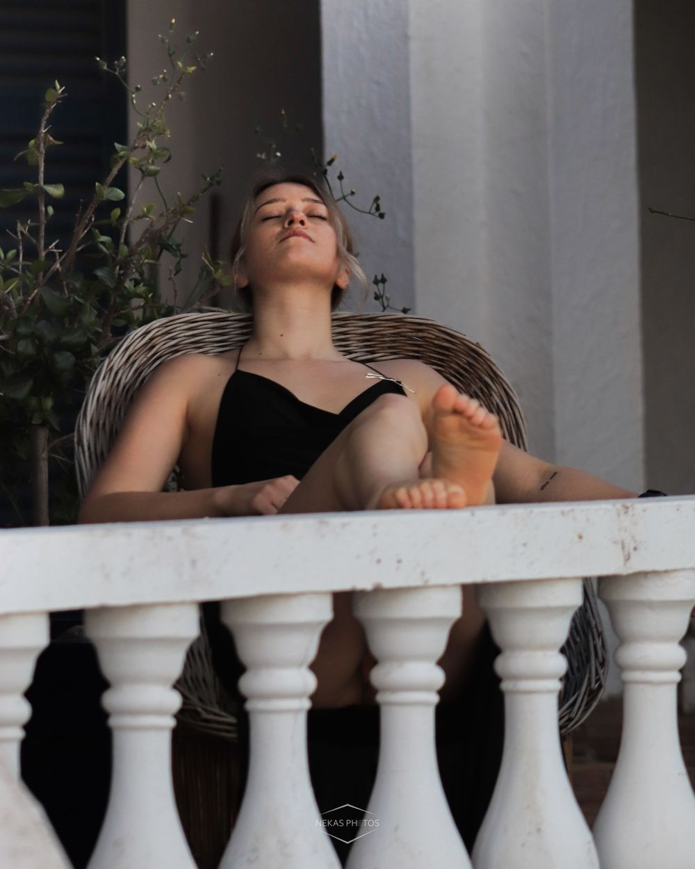 Woman relaxing on a balcony chair, feet up on white balustrade, eyes closed, enjoying sunlight.