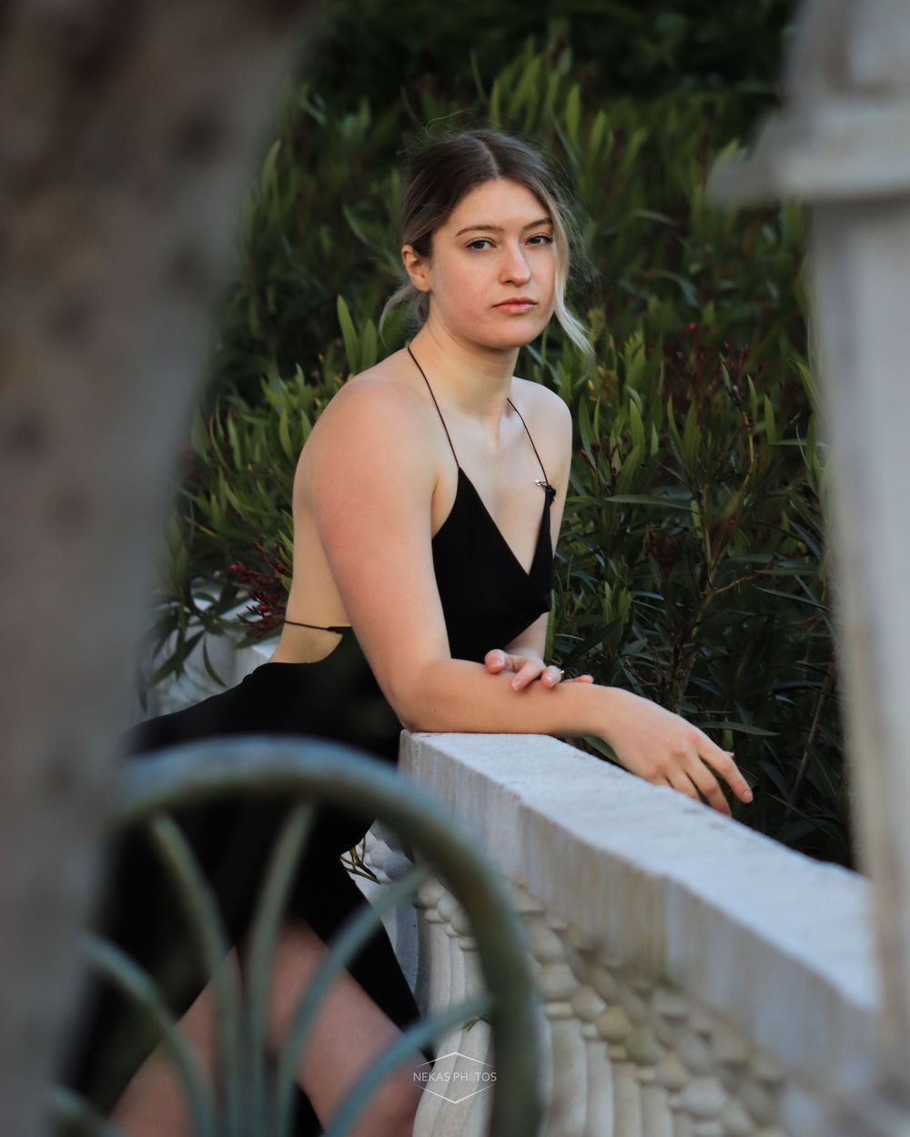 Young woman in black dress leaning on white railing outdoors, surrounded by lush green foliage.