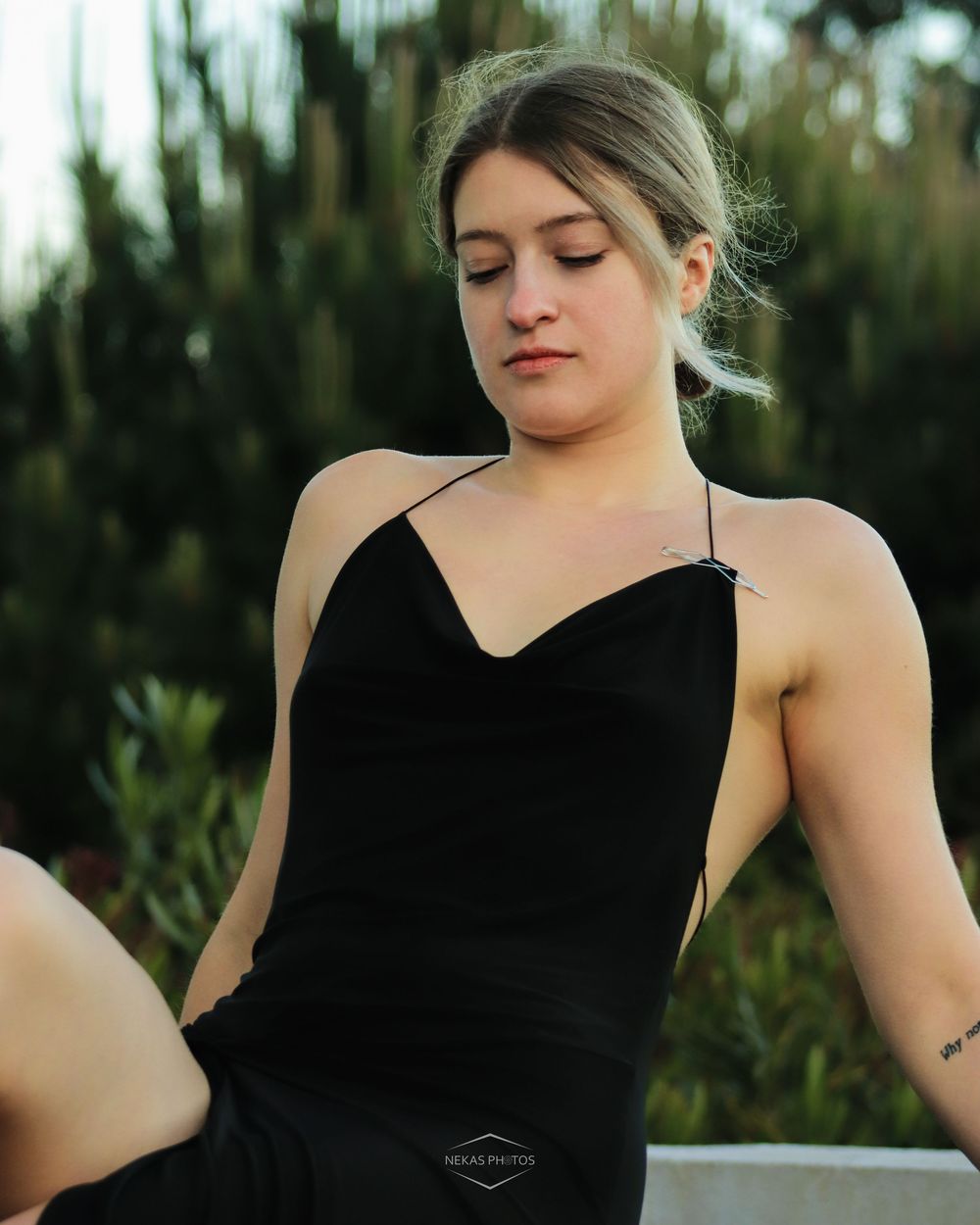Young woman in black spaghetti strap dress sitting outdoors with green foliage background, looking down pensively.